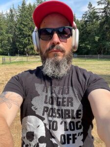 A recent photograph of the author, standing in a field with a beard, wearing sunglasses, a cap, large headphones and a shirt by dashare.zone reading IT IS NO LONGER POSSIBLE TO 'LOG OFF'